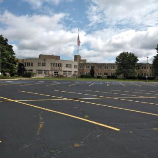 A view of a school building with a vast parking lot under a partly cloudy sky.