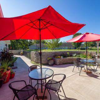 A sunny outdoor patio featuring red umbrellas, tables, and chairs in a garden setting.