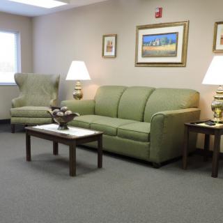 A cozy waiting room with a green couch, armchair, and coffee table adorned with decorative items, illuminated by two lamps and complemented by framed artwork on the walls.