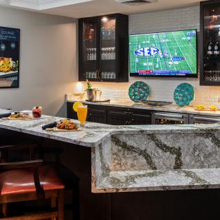 A modern home bar setup featuring a granite countertop with seating, decorated with vibrant accents and a television displaying a football game.