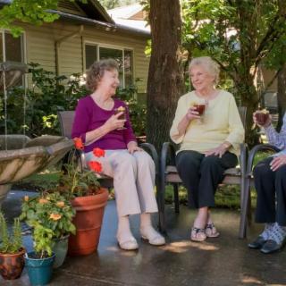 Three elderly women enjoying a conversation and drinks in a garden setting.