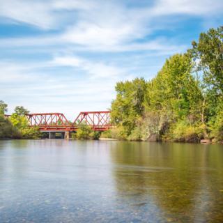 A serene view of a red bridge spanning over a calm river, surrounded by lush greenery under a bright blue sky.