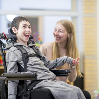 A caregiver and a child in a wheelchair share a joyful moment indoors.