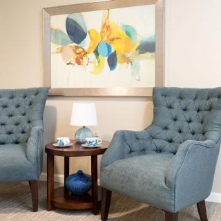 A cozy living room corner with two tufted blue armchairs, a side table with a lamp and decorative items, and a colorful abstract painting on the wall.