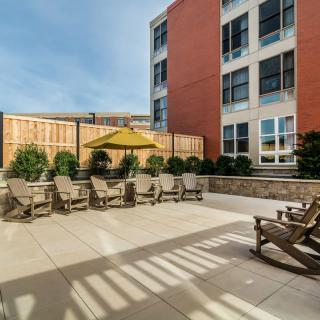 An outdoor patio area with wooden chairs and a yellow umbrella, adjacent to a modern building.