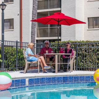 A group of people sitting by the pool under a red umbrella, enjoying a sunny day.