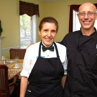 Two restaurant staff members dressed in uniforms and aprons, smiling indoors.
