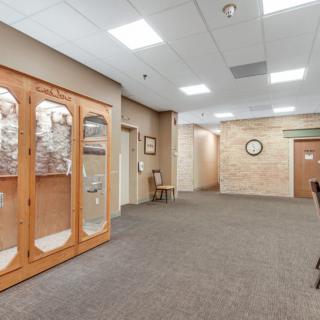 A carpeted hallway with a wooden cabinet, clock on the wall, and seating area under bright ceiling lights.