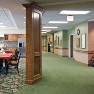 A spacious hallway in a nursing home with a dining area and kitchen, featuring green carpet and wood paneling.