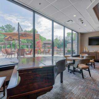 A modern lounge with a grand piano and seating area, featuring large windows overlooking a patio with umbrellas.
