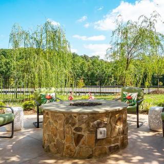 An inviting outdoor patio featuring a stone fire pit surrounded by chairs, set against a backdrop of lush greenery and trees.