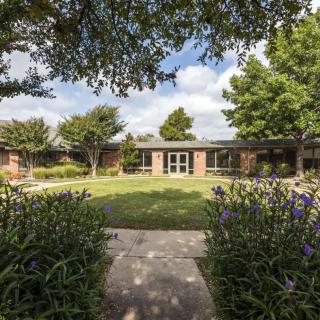 A tranquil courtyard with a pathway leading to a brick building surrounded by lush greenery and trees.