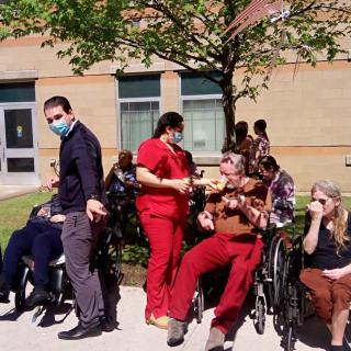 A group of people, including healthcare workers and individuals in wheelchairs, gathered outdoors on a sunny day.