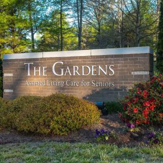 The entrance sign to The Gardens, surrounded by vibrant flowers and lush greenery.