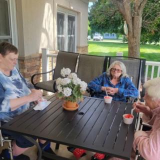 Three elderly women enjoy a conversation on a sunny patio, surrounded by flowers.