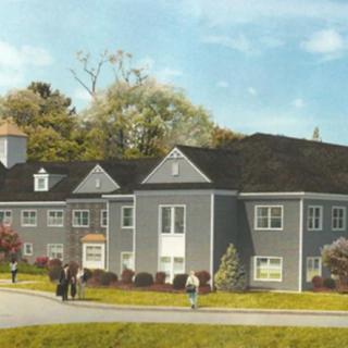 A residential building surrounded by trees and grass with people walking nearby.