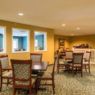 A cozy dining area with tables and chairs, featuring warm lighting and carpeted flooring.