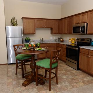 A cozy kitchen with wooden cabinets, stainless steel appliances, and a small dining area featuring a round table with green-cushioned chairs.