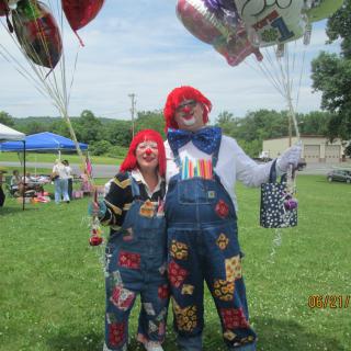 Two clowns in colorful overalls holding balloons at an outdoor event.