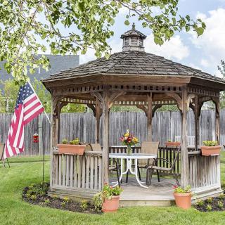 A wooden gazebo in a garden setting with a table and chairs, surrounded by flower pots and an American flag.