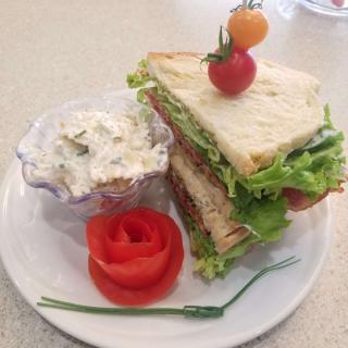 A stacked sandwich with greens and cherry tomatoes, served alongside a creamy salad and garnished with a rose-shaped tomato.