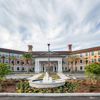 A grand building with a central fountain, surrounded by landscaped greenery and a cloudy sky.