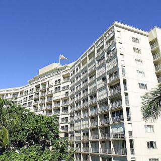 A large residential apartment building with balconies, surrounded by lush palm trees under a clear blue sky.