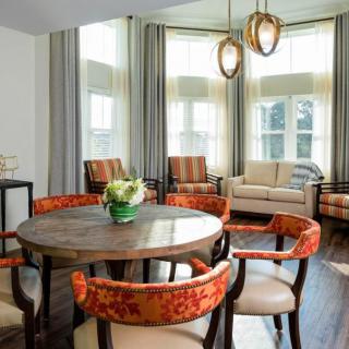 A well-decorated living room featuring a round dining table with orange upholstered chairs, large windows with curtains, and modern lighting.