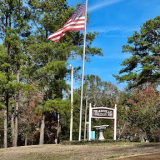 A welcoming sign stands tall with an American flag waving in the breeze, surrounded by lush trees and a clear blue sky.