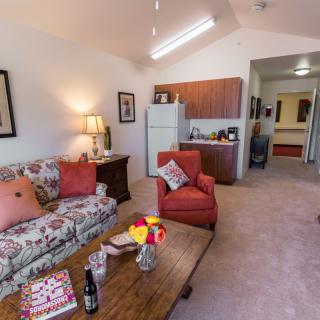 A cozy living room with a floral sofa, red armchair, and a wooden coffee table adorned with flowers and decorative items. The kitchenette in the background adds to the homely atmosphere.