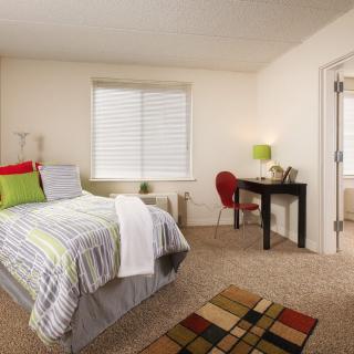 A cozy bedroom with a neatly made bed, a desk with a lamp, and a small carpet on the floor.