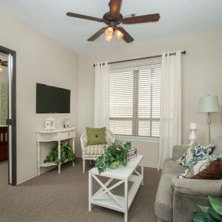 A cozy living room with beige walls, featuring a comfortable sofa, a white coffee table adorned with plants, and a wall-mounted TV, illuminated by natural light streaming through the window.