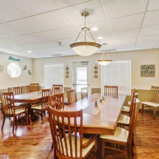 A spacious dining room with wooden tables and chairs, illuminated by overhead chandeliers, featuring natural light from large windows.