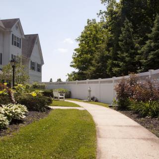 A scenic garden walkway bordered by flowers and a white fence with a house in the background.