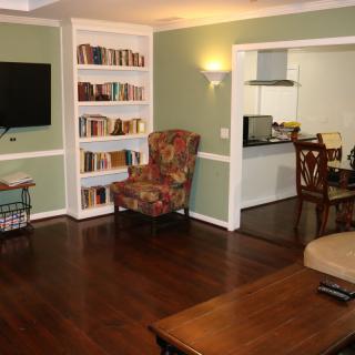 A cozy living room with hardwood floors, featuring a bookshelf, armchair, and wall-mounted TV, with an open view to the kitchen.