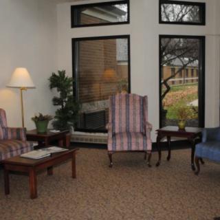 A cozy living room with comfortable chairs, a lamp, and large windows overlooking a garden.