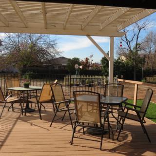 A sunny patio with tables and chairs under a pergola, overlooking a suburban yard.