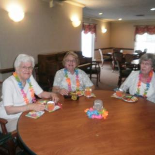 Three elderly women enjoying a meal together at a dining table, adorned with colorful leis.