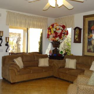A cozy living room with brown sofas, decorative pillows, and a large floral arrangement. The space is accented by wall art and lit by a ceiling fan.