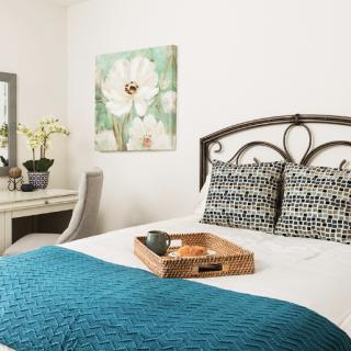A cozy bedroom featuring a bed with patterned pillows, a teal blanket, and a decorative tray. The room is complemented by a floral painting and a mirror on the wall.