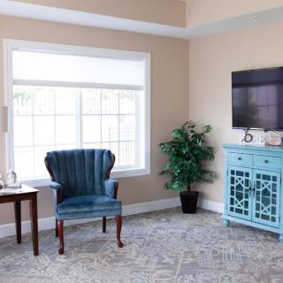 A cozy living room with a blue wingback chair, large window, and a television mounted above a turquoise cabinet.
