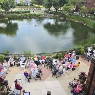A group of people gather outdoors by a pond, sitting on chairs and enjoying an event.