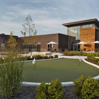 A modern building with a landscaped outdoor area featuring a small golf putting green and patio umbrellas.