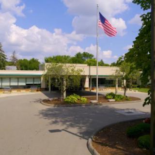 A school building with an American flag in front, surrounded by trees and a driveway.