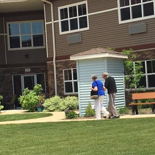 A caregiver assists an elderly person outside a residential building with a garden.