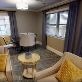 A cozy living room with beige chairs, a round table, lamp, and large windows with curtains.
