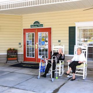 Two elderly individuals relax in rocking chairs on the front porch of a retirement home.
