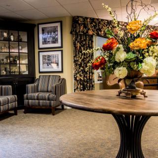 A cozy lounge area with elegant decor, featuring armchairs, a floral arrangement on a table, and framed pictures on the wall.