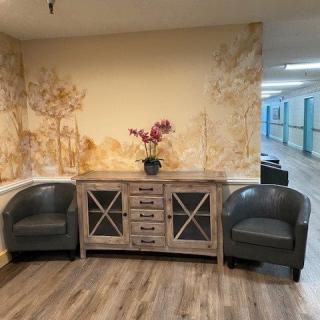 A cozy hallway corner featuring two leather chairs, a wooden cabinet with decorative glass doors, and a potted orchid on top. The backdrop is a mural-style wall art adding warmth to the ambiance.