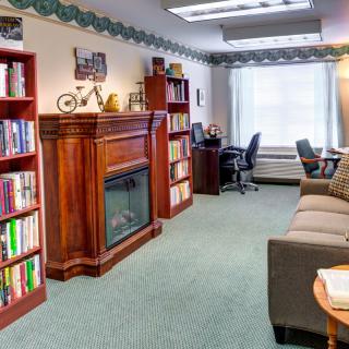 A cozy office space featuring bookshelves, a fireplace, desk, and couch with natural light streaming in through a window.